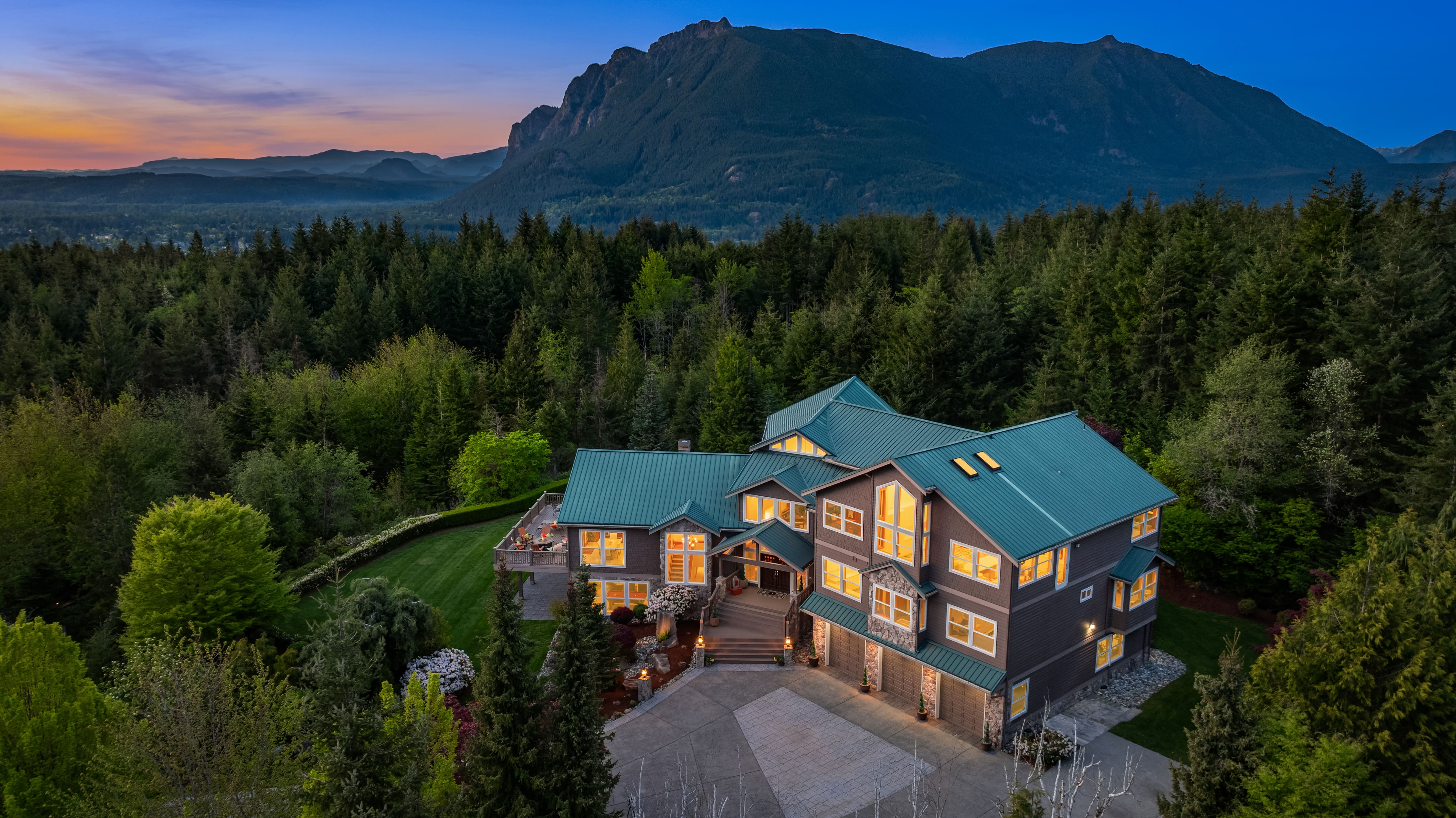 Stunning exterior view of a luxury home at twilight, surrounded by lush greenery and mountains, located in the Pacific Northwest. The property features a modern design with large windows and a distinctive metal roof, creating an inviting and serene atmosphere.