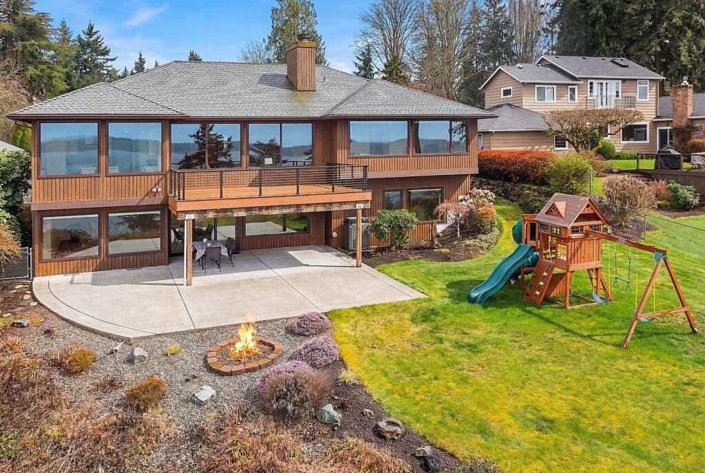 Modern exterior of a spacious home with large windows, featuring a patio area with a fire pit and a children's playset on a lush green lawn, located in the Pacific Northwest.