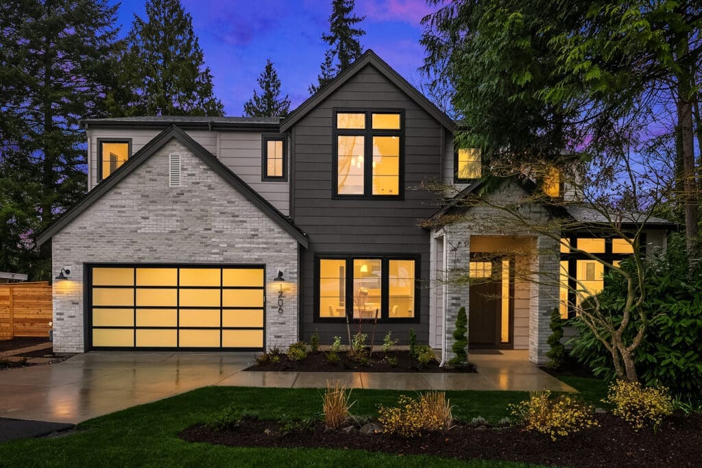 Modern exterior of a stylish home in the Pacific Northwest, featuring a blend of brick and dark siding, illuminated windows, and landscaped yard at twilight.