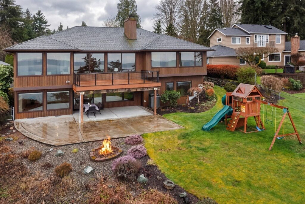 Spacious modern exterior of a waterfront home in the Pacific Northwest, featuring large windows, a wraparound deck, a fire pit, and a children's playset in the lush backyard.